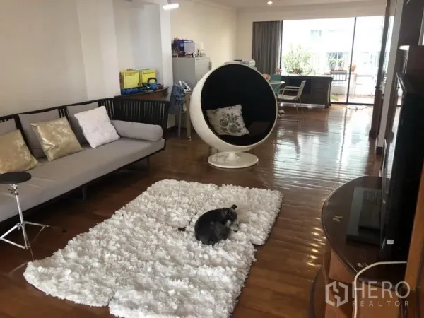 Condominium for sale in Watthana, Bangkok - Spacious living area with polished wood floors, modern egg chair, sofa, TV unit and sliding doors to a balcony.