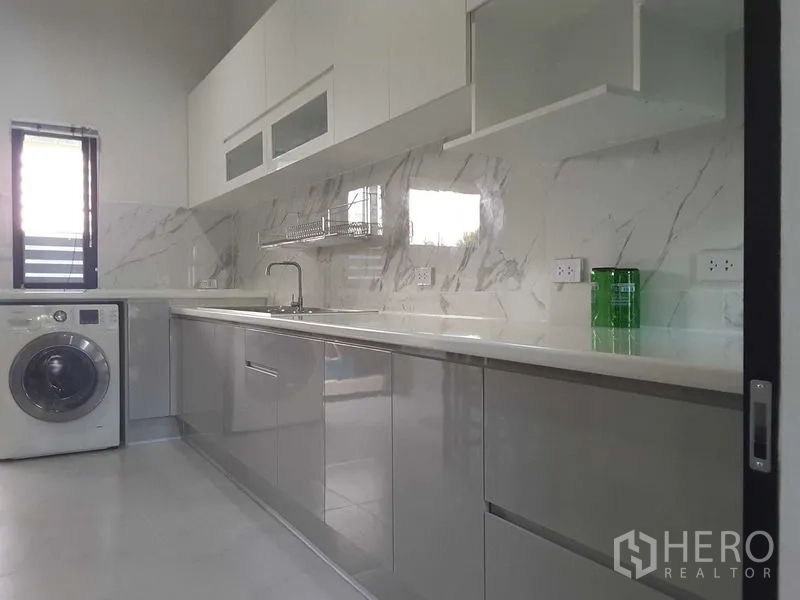 Detached House for rent or sale in Lam Luk Ka, Pathum Thani - Bright white kitchen with marble backsplash, long countertop, upper cabinets and a washing machine by the window.