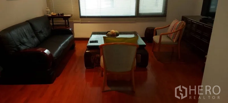 Condominium for rent in Sathon, Bangkok - Cozy living room with black leather sofa, glass coffee table, wood floors and large window blinds.
