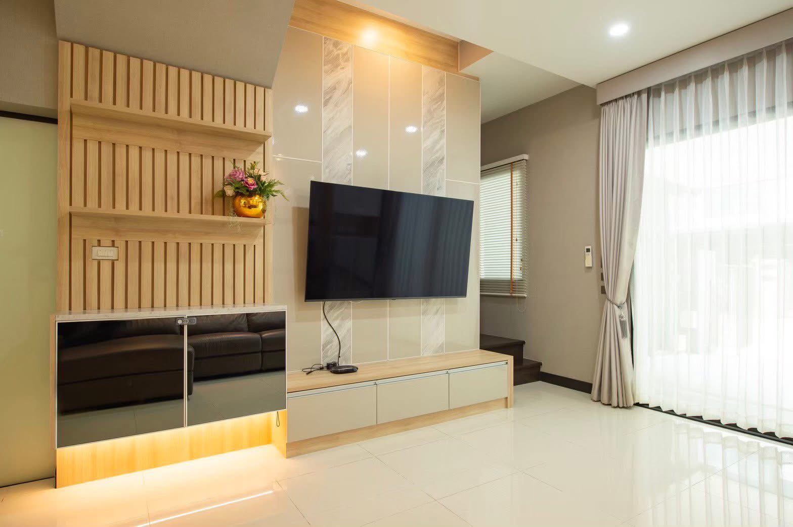 Townhouse for rent or sale in Lat Phrao, Bangkok - TV wall with floating cabinets, marble accents and sliding drapes beside the stairway.
