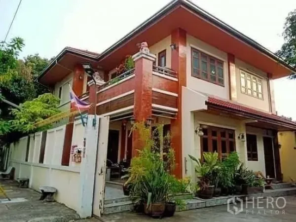 Detached House for sale in Bang Na, Bangkok - Front exterior of the two-storey house with balcony, potted greenery and driveway.