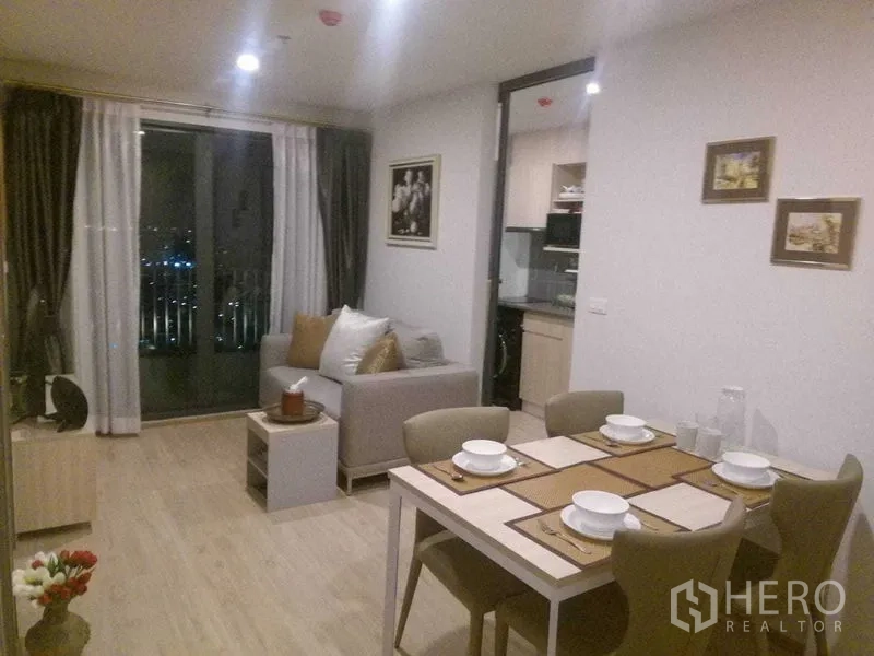 公寓 出租在Bang Na, Bangkok - 夜间开放式客厅，四人餐桌、沙发，厨房旁是通向阳台的推拉门。