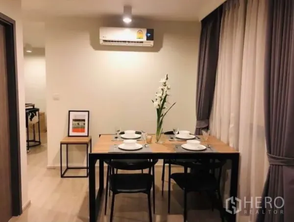 Condominium for rent or sale in Pathum Wan, Bangkok - Dining area with a wooden table set for four beneath an air conditioner and sheer drapes.