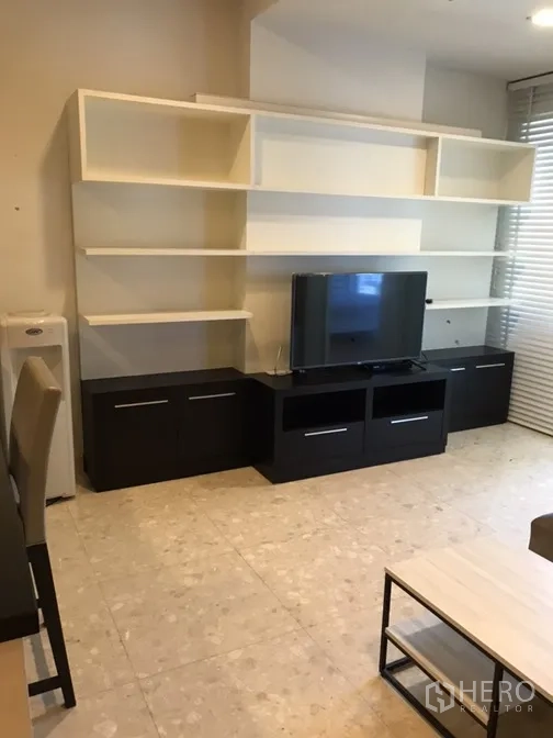 Condominium for rent in Khlong Toei, Bangkok - Living room media wall with open white shelving, black TV console and window blinds.