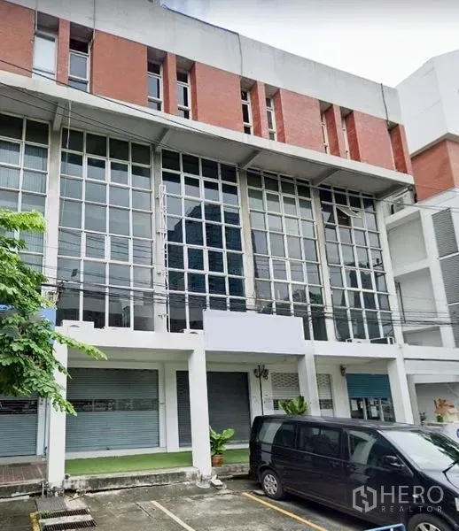店屋 出租在Watthana, Bangkok - 瓦他那区四层商铺外立面，落地玻璃窗、卷闸门与门前停车位，临近Ekkamai轻轨站