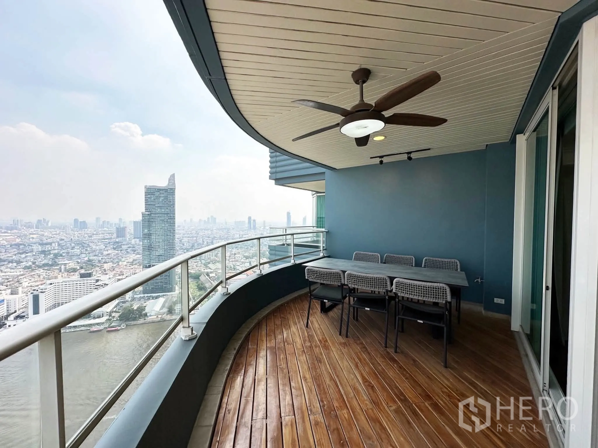 คอนโด for sale in Khlong San, Bangkok - ระเบียงโค้งปูพื้นไม้พร้อมพัดลมเพดานและชุดโต๊ะทานอาหาร มองเห็นวิวแม่น้ำและเมืองกว้างไกล