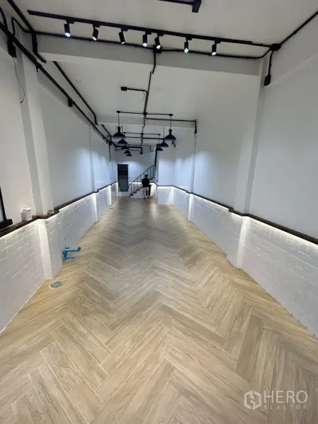 Shophouse for rent in Khlong Toei, Bangkok - Long retail hall with herringbone wood-look flooring, white tile wainscoting, track lights, and rear stairs.