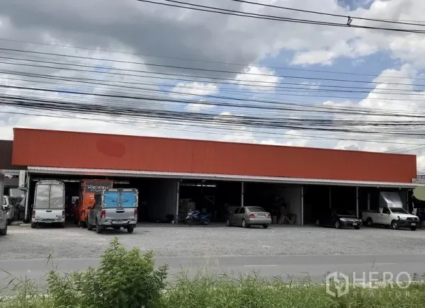 Warehouse for rent in Pathum Thani - Wide view of a single‑storey warehouse with orange cladding, open loading bays and a gravel forecourt with parked trucks and cars.