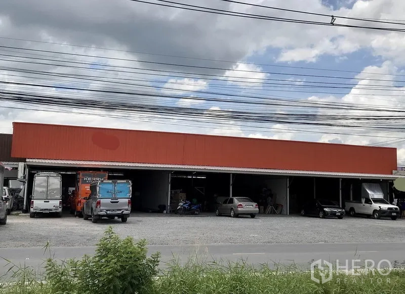 Warehouse for rent in Pathum Thani - Wide view of a single‑storey warehouse with orange cladding, open loading bays and a gravel forecourt with parked trucks and cars.