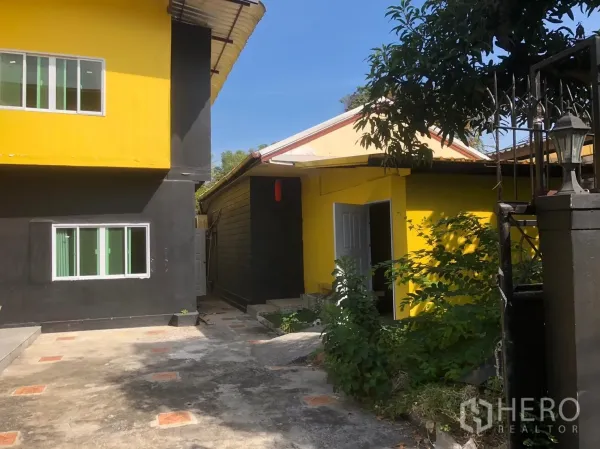 Detached House for rent in Khlong Toei, Bangkok - Side courtyard between the main house and a yellow annex with sliding windows and greenery along a paved path.