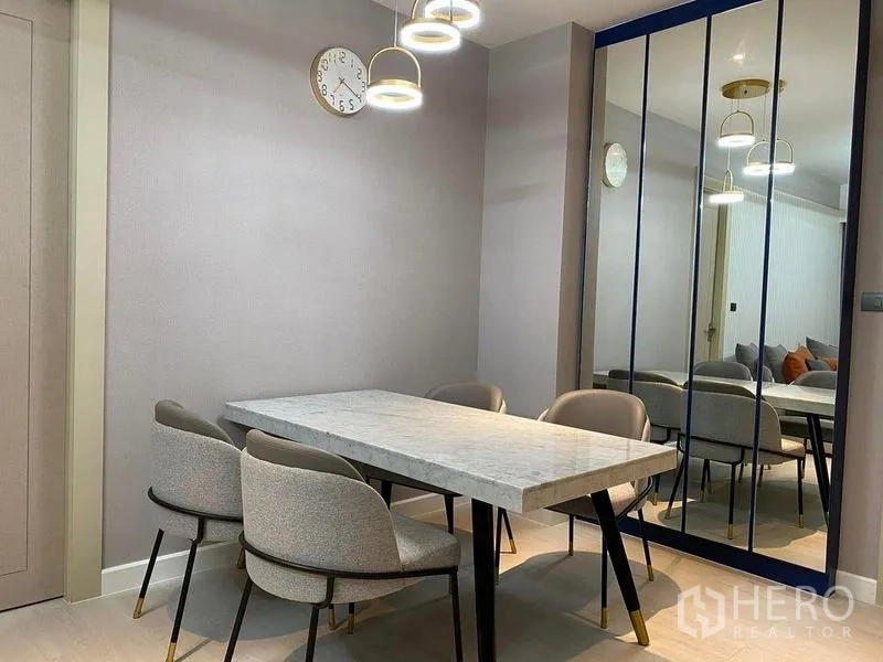 Condominium for sale in Huai Khwang, Bangkok - Close-up of dining area showing marble tabletop, soft gray chairs and full-height mirrored wall with ring pendants.