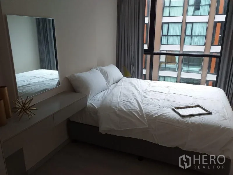Condominium for rent in Khlong Toei, Bangkok - Cozy bedroom with white bedding, dressing console, and floor-to-ceiling window overlooking the neighboring building.