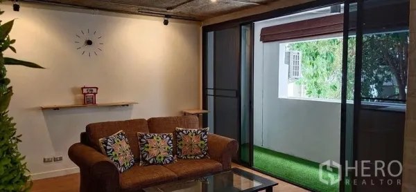 Condominium for sale in Watthana, Bangkok - Cozy living room with brown sofa, glass coffee table and sliding doors opening to a balcony with artificial grass.