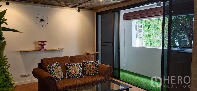 Condominium for sale in Watthana, Bangkok - Cozy living room with brown sofa, glass coffee table and sliding doors opening to a balcony with artificial grass.