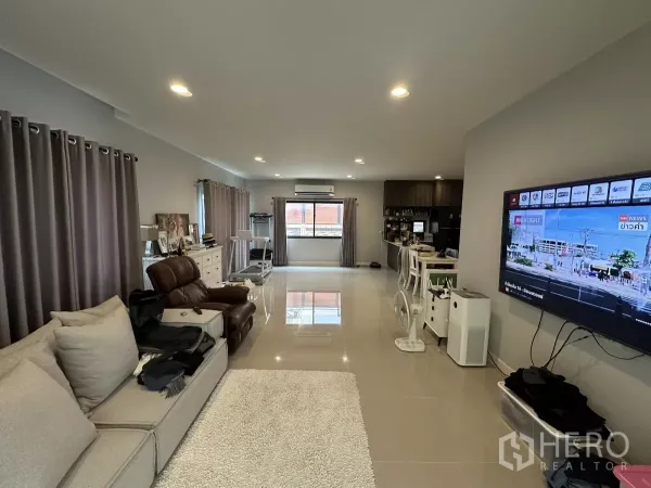 Detached House for rent or sale in Lam Luk Ka, Pathum Thani - Spacious open-plan living room with sectional sofa, wall-mounted TV, recessed lighting and glossy tile floors.