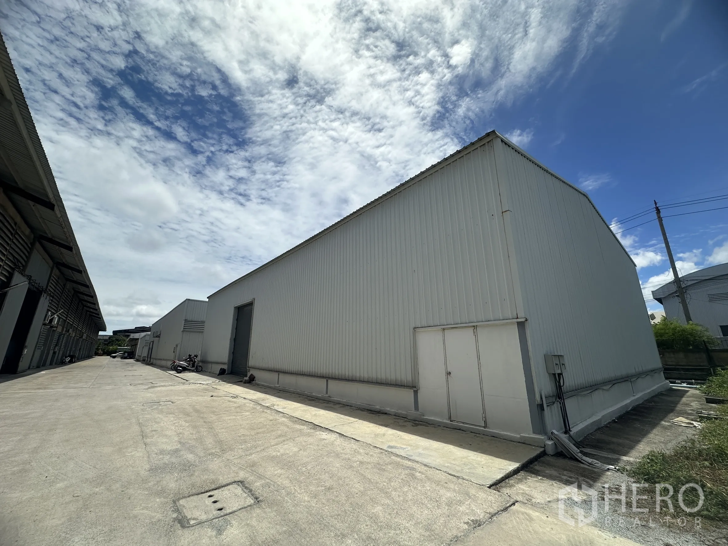 Factory for rent in Mueang Samut Prakan, Samut Prakan - Side view of the warehouse-style factory along a wide driveway with utility doors and power box.