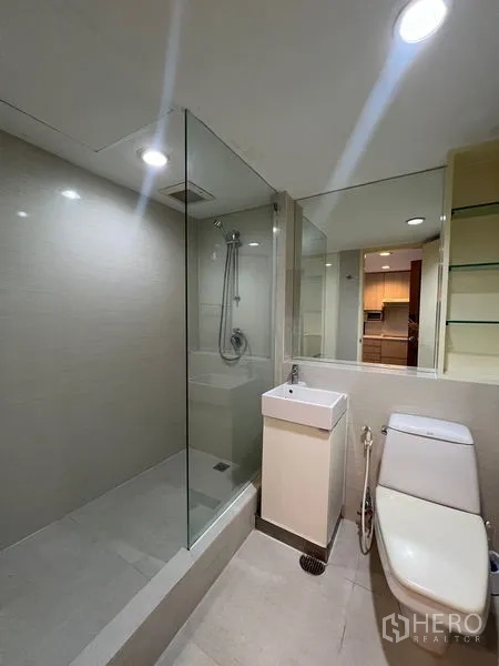 Condominium for rent or sale in Khlong Toei, Bangkok - Bathroom with glass-enclosed shower, vanity and toilet under recessed lights.