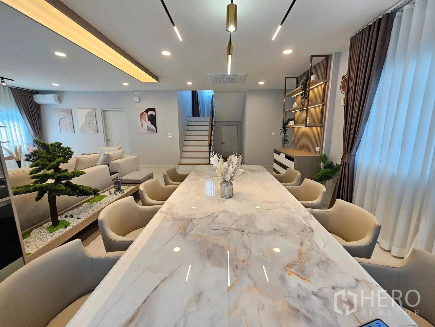 Detached House for rent or sale in Bang Phli, Samut Prakan - Dining area with long marble table, upholstered chairs, bonsai planter and stairs beyond.