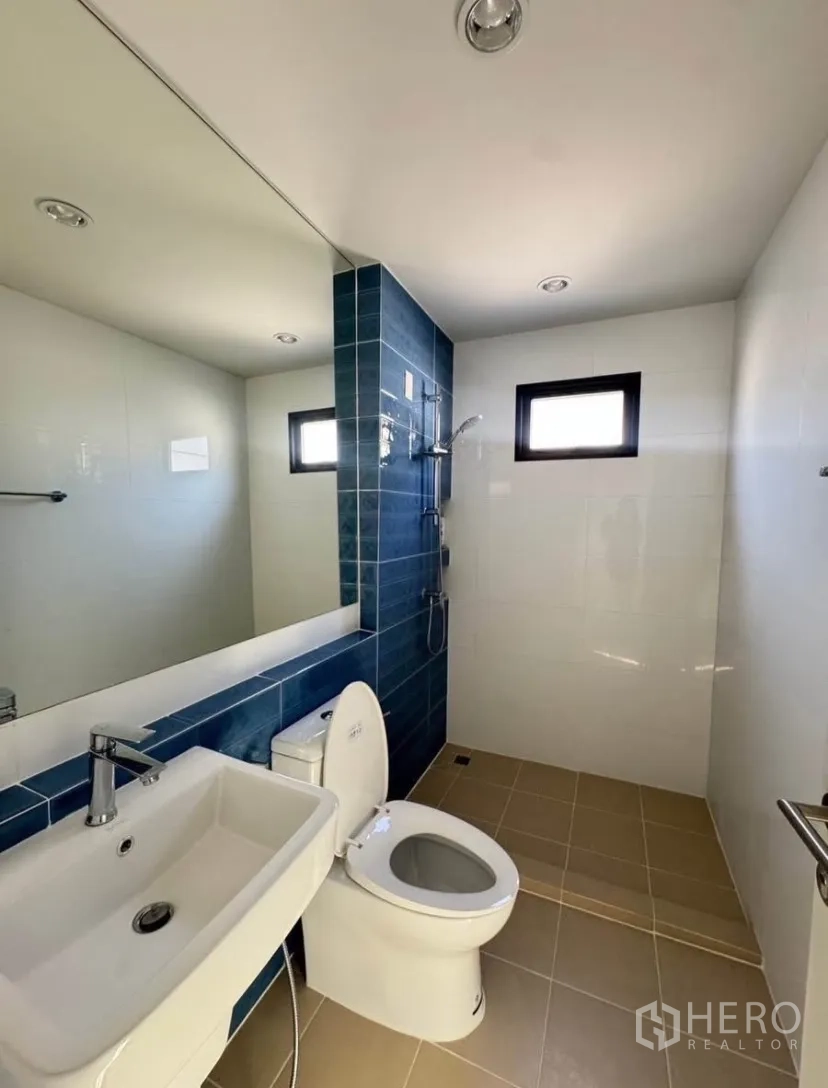 Detached House for rent or sale in Lat Krabang, Bangkok - Modern bathroom with blue accent tiles, wide mirror, wall-mounted sink, toilet, and walk-in shower with window.