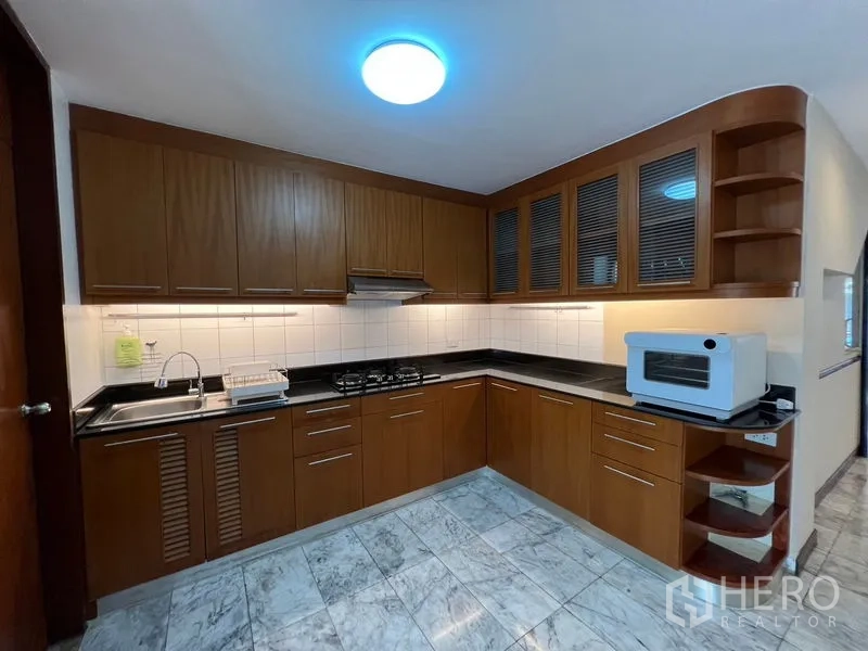 Townhouse for rent or sale in Suan Luang, Bangkok - L-shaped kitchen with wood cabinets, black countertops and a gas stove.