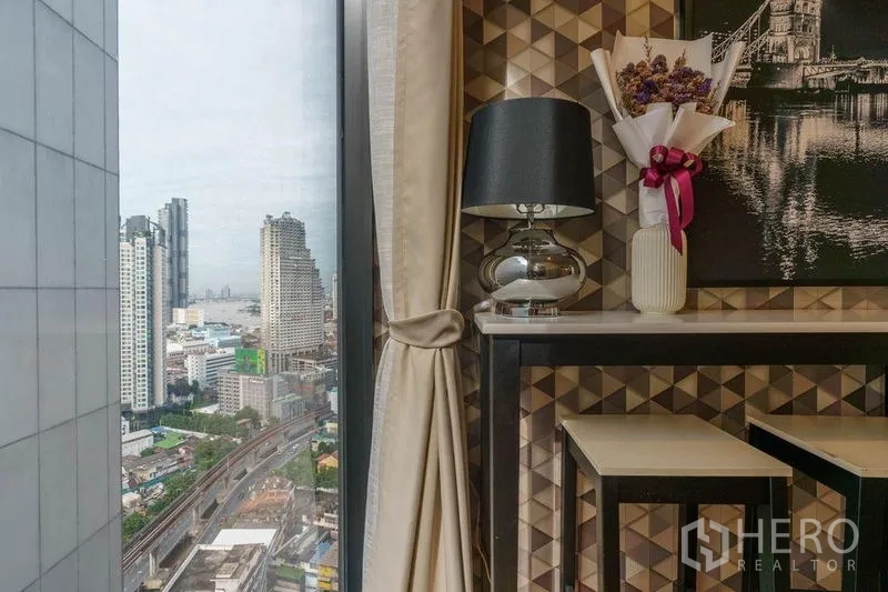 公寓 出租或出售在Bang Rak, Bangkok - 窗角休闲区，台灯与装饰画，窗外可见高楼与轻轨轨道。