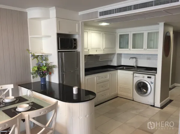 Condominium for rent in Khlong Toei, Bangkok - U-shaped white kitchen with black countertops, sink, fridge, microwave, and in-unit washing machine beside dining area.