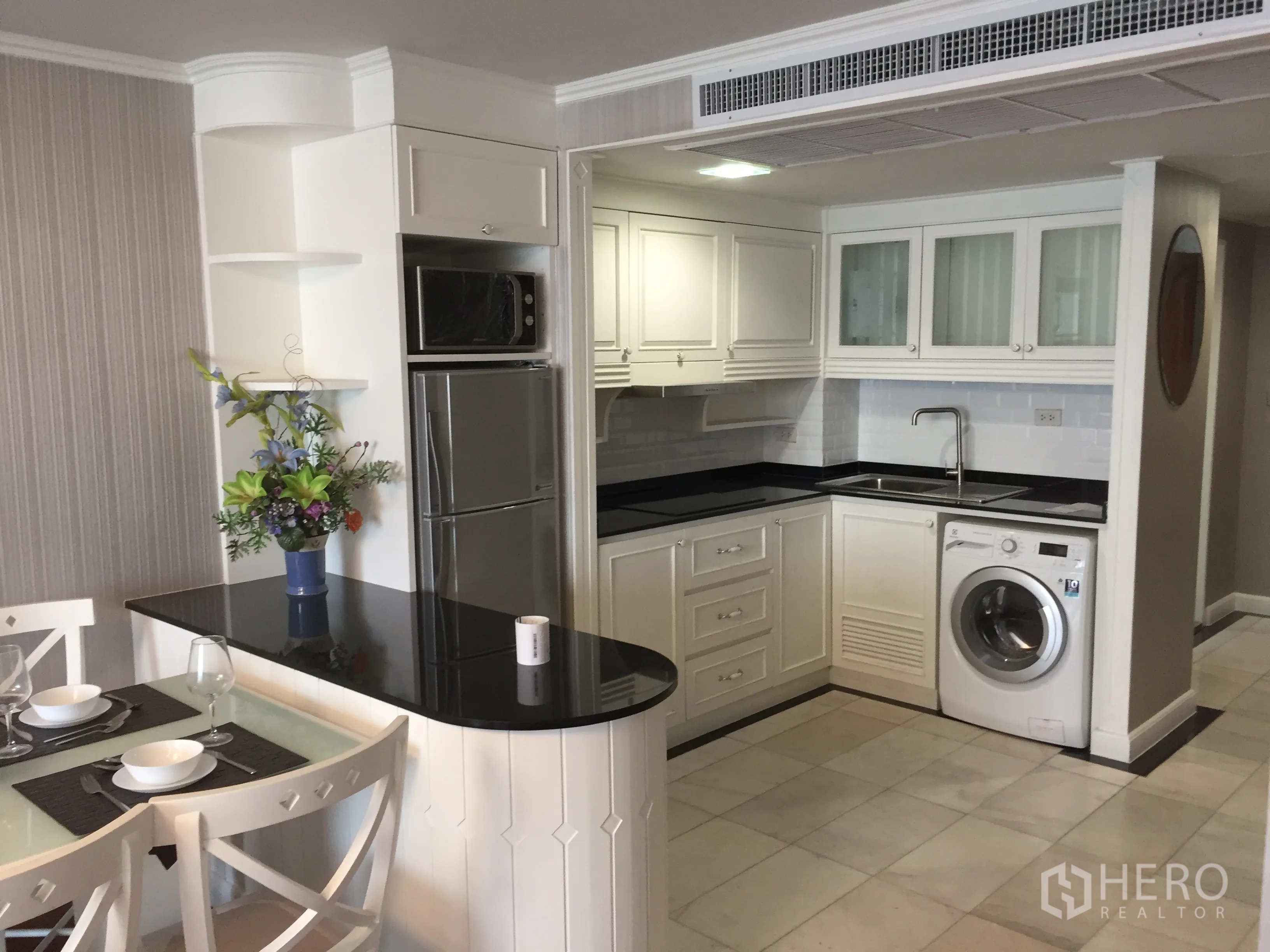 Condominium for rent in Khlong Toei, Bangkok - U-shaped white kitchen with black countertops, sink, fridge, microwave, and in-unit washing machine beside dining area.