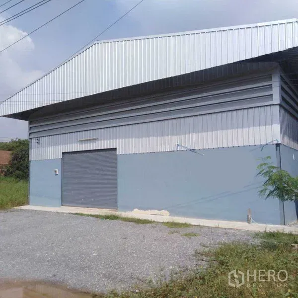 Warehouse for rent in Bang Yai, Nonthaburi - Side exterior of a metal-clad warehouse with a roller shutter door and gravel parking area.
