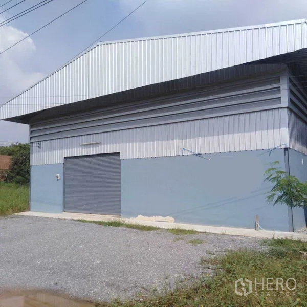 Warehouse for rent in Bang Yai, Nonthaburi - Side exterior of a metal-clad warehouse with a roller shutter door and gravel parking area.