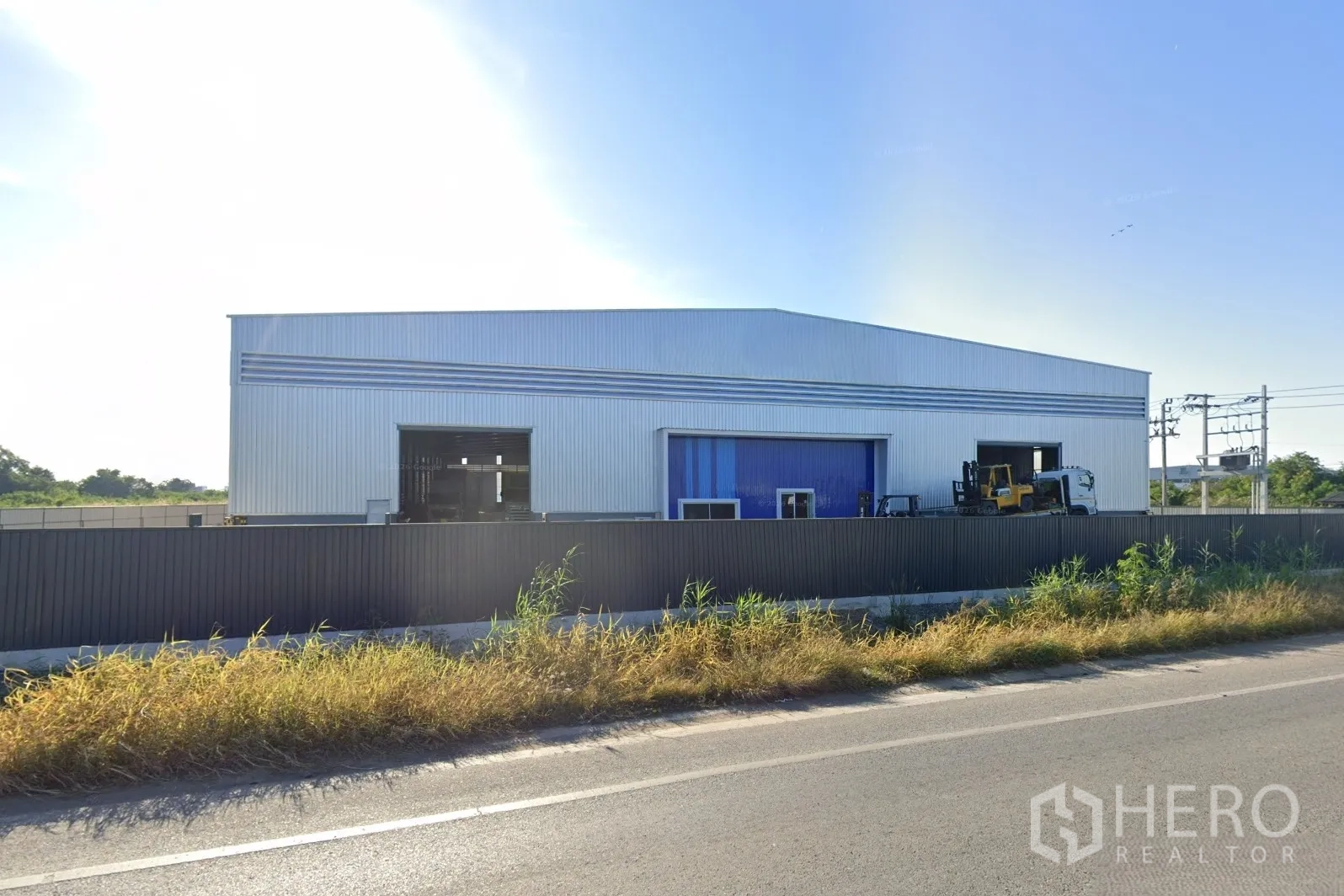 Factory for rent in Bang Bo, Samut Prakan - Modern metal-clad factory with multiple wide roller shutter doors, secure perimeter fencing, and road frontage under a clear sky.