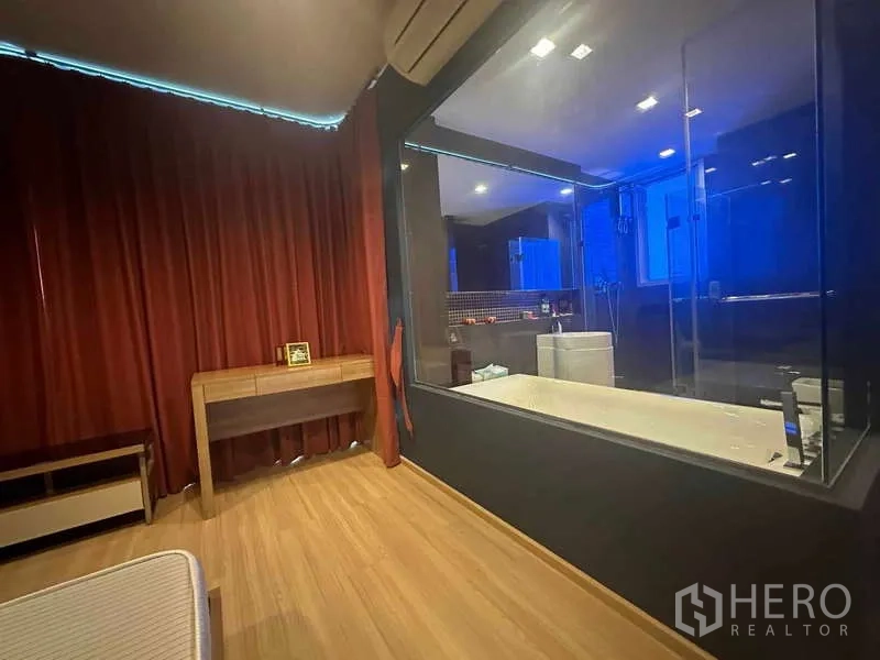 Condominium for sale in Phaya Thai, Bangkok - Bedroom with large glass partition looking into a bathroom with soaking tub and red blackout curtains.