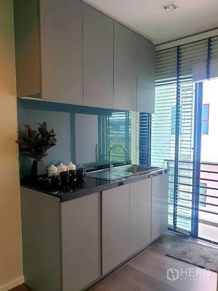 Home Office for rent in Bang Phli, Samut Prakan - Compact kitchenette with grey cabinetry, black countertop, sink and a sliding glass door with blinds.