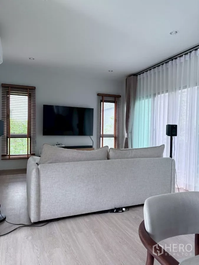 Detached House for rent in Lat Krabang, Bangkok - Bright living room with grey sofa, wall-mounted TV and sheer curtains.