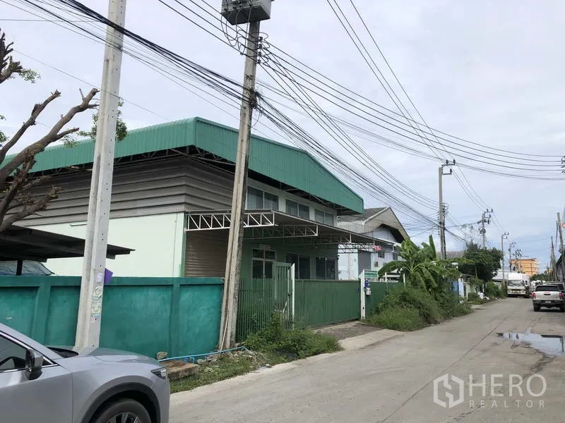 Factory for rent or sale in Samut Prakan - Corner view of the factory exterior along the access road with overhead power lines and on-street parking.