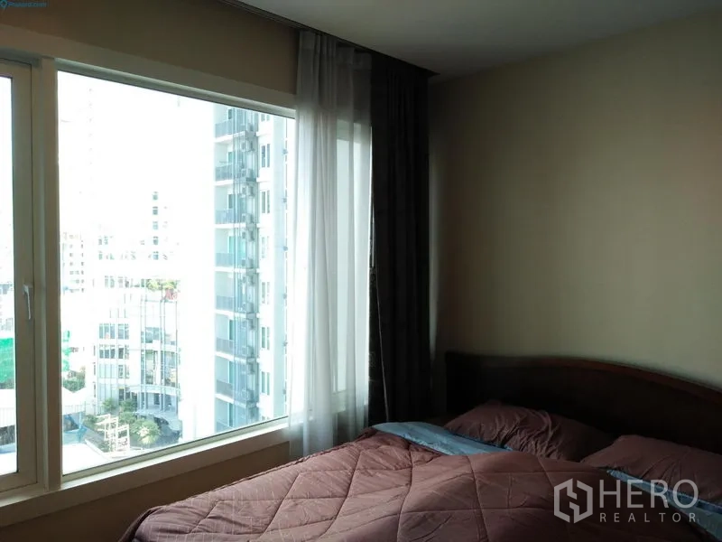 Condominium for rent in Khlong Toei, Bangkok - Bedroom corner with large window offering city views through sheer curtains beside the bed.