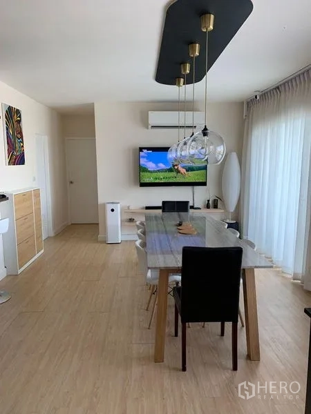 公寓 出租在Bang Na, Bangkok - 从餐区望向电视墙，现代吊灯、浅色木地板与推拉玻璃门。