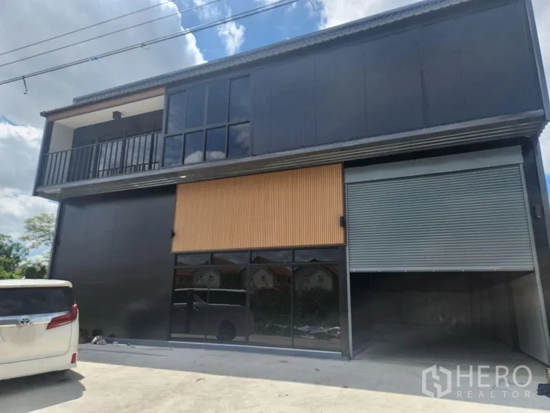 Warehouse for rent or sale in Lam Luk Ka, Pathum Thani - Angled exterior view of the modern warehouse showing a glass storefront, balcony and a wide roller shutter entrance.