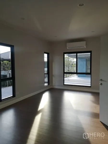 Detached House for sale in Lam Luk Ka, Pathum Thani - Sunlit bedroom with corner windows, air conditioner and wood-look floors.