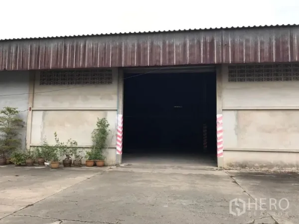 Warehouse for rent in Mueang Nakhon Pathom, Nakhon Pathom - Warehouse front facade with a large entrance and concrete yard with potted plants.