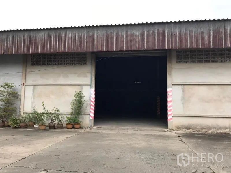 Warehouse for rent in Mueang Nakhon Pathom, Nakhon Pathom - Warehouse front facade with a large entrance and concrete yard with potted plants.