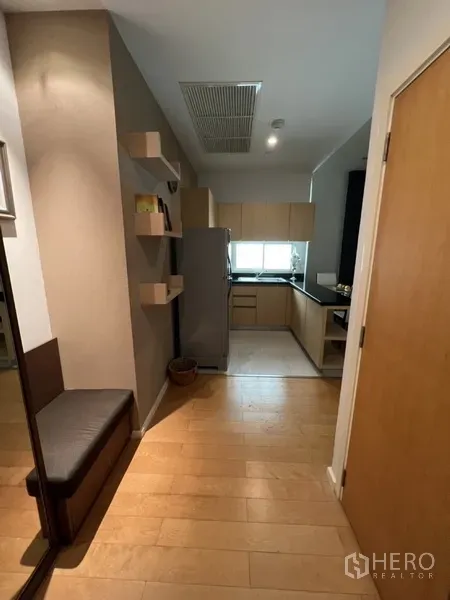 Condominium for rent in Chatuchak, Bangkok - Entry hall with wall shelves leading to an L-shaped kitchen with window and light wood cabinetry.