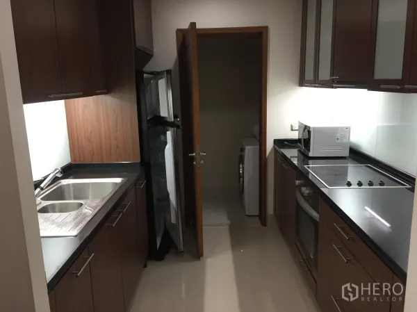 Condominium for rent in Sathon, Bangkok - Galley kitchen with wood cabinets, black stone counters, electric hob and oven, fridge, and a utility area with washing machine.