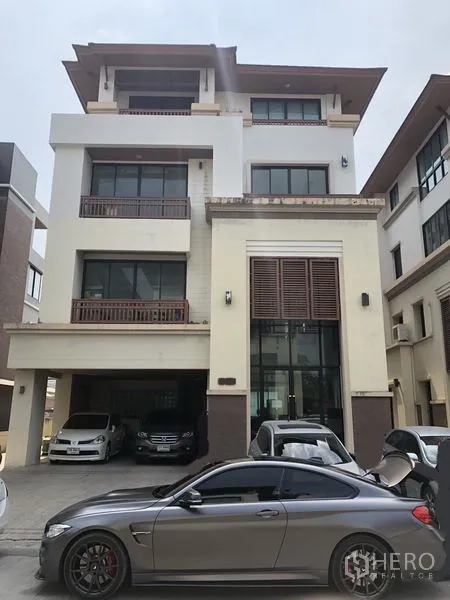 带办公室的家 出租在Wang Thonglang, Bangkok - 多层居家办公楼外观，挑高玻璃入口、阳台与有顶停车位，前方停有多辆车。