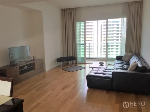 Condominium for rent in Khlong Toei, Bangkok - Bright living room with black leather sectional, wood floors, TV console, and sliding doors to a balcony with city view.