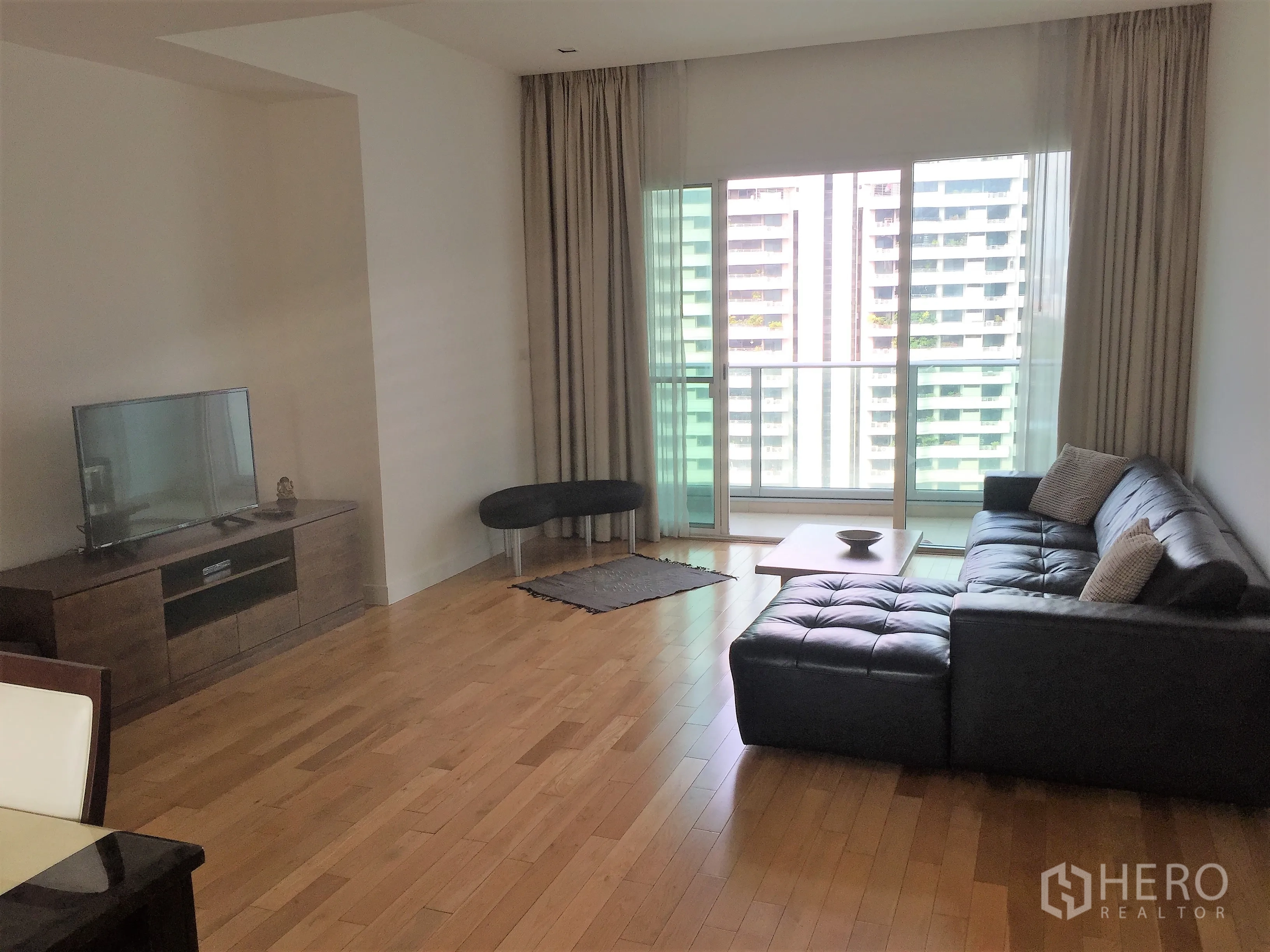 Condominium for rent in Khlong Toei, Bangkok - Bright living room with black leather sectional, wood floors, TV console, and sliding doors to a balcony with city view.