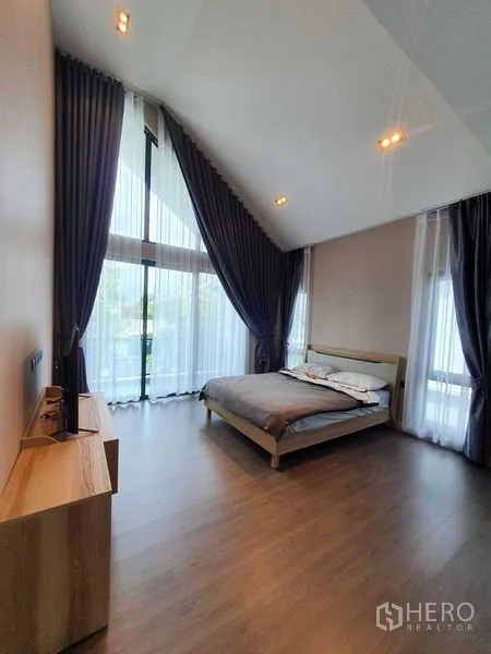 Detached House for rent in Khlong Luang, Pathum Thani - Spacious bedroom featuring a vaulted ceiling and floor-to-ceiling windows with dark curtains.