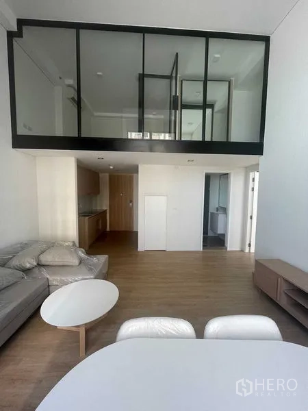 Condominium for rent in Khlong Toei, Bangkok - Open-plan living and dining with sofa and tables, beneath a glass-walled loft bedroom.