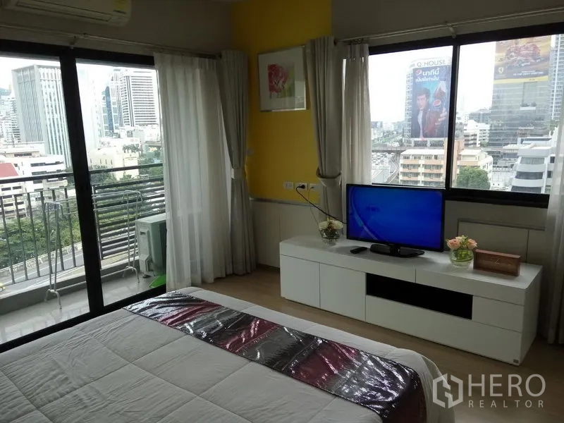 公寓 出租或出售在Pathum Wan, Bangkok - 转角卧室，落地窗与阳台可观城市天际线，白色柜上摆放电视。