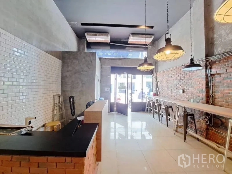 零售空间 出租在Watthana, Bangkok - 工业风商铺内景，高挑层空间，砖与瓷砖墙面，吊灯、长吧台与临街玻璃门面。