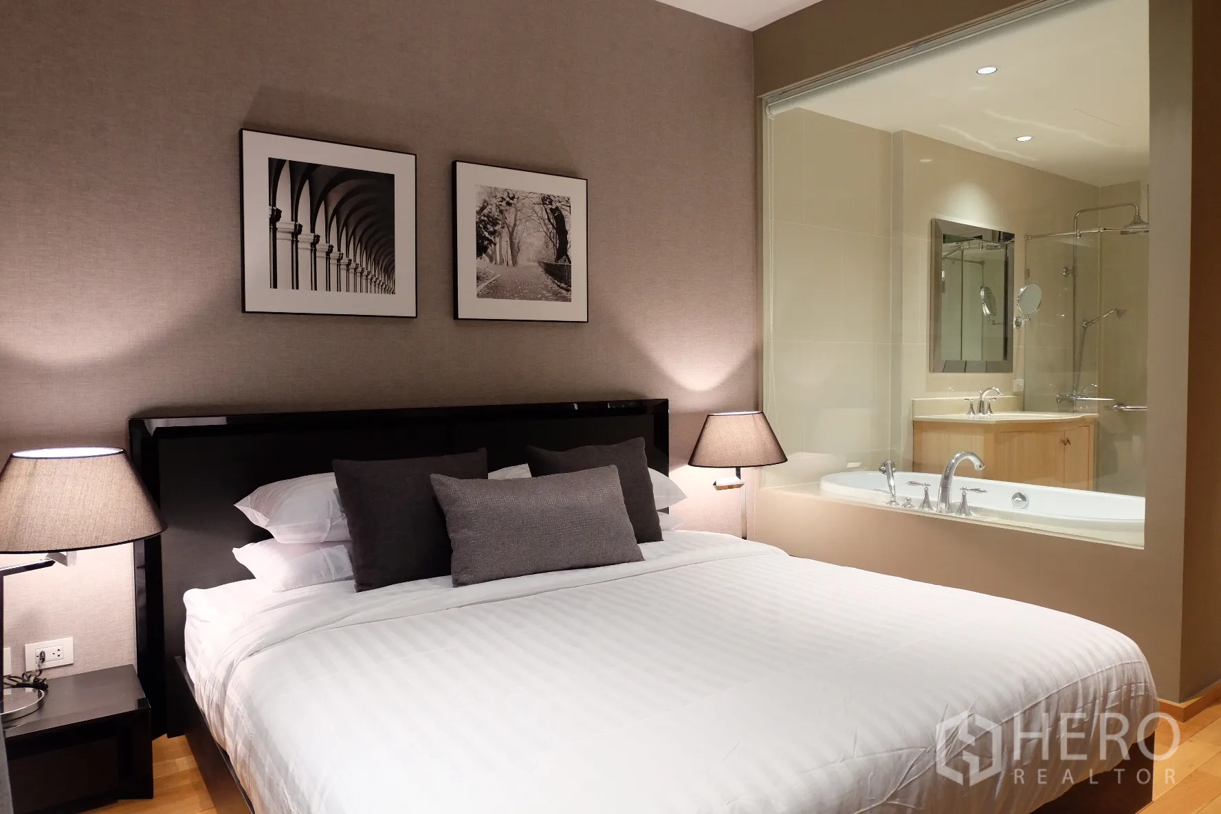 公寓 出租在Sathon, Bangkok - 主卧配特大床与床头灯，墙面装饰画，玻璃窗可见带浴缸的套卫。